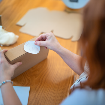 Eine Person hält ein braunes Paket mit einem Aufkleber in der Hand, während sie an einem Holztisch sitzt und arbeitet.