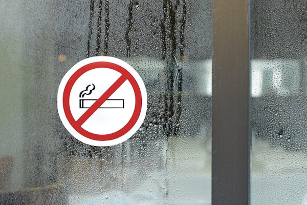 Ein rundes Schild mit einem durchgestrichenen Zigaretten-Symbol, das ein Rauchverbot signalisiert, hängt an einer beschlagenen Glasfläche.