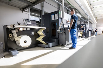 Ein Arbeiter bedient eine moderne Druckmaschine in einer Produktionshalle, während eine Rolle Papier bereitgestellt wird.
