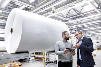 Zwei Männer in einer Fabrik diskutieren über ein Tablet, während im Hintergrund eine große Rolle Papier steht.