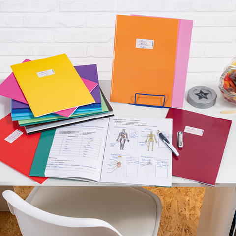 Bunte Ordner und Unterlagen auf einem Schreibtisch, daneben ein Glas mit Süßigkeiten und Bürobedarf wie einen Hefter und einen Stift.