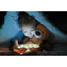 Ein Kind sitzt im Bett, hält einen leuchtenden Gegenstand in den Händen und umarmt einen Teddy, während es unter einer Decke liegt.