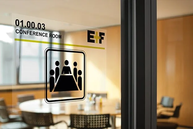 Eine Visitenkarte und ein Umschlag liegen auf einem Tisch, daneben steht ein Glastürschild mit einem Symbol für einen Konferenzraum und drei Parfümflaschen.