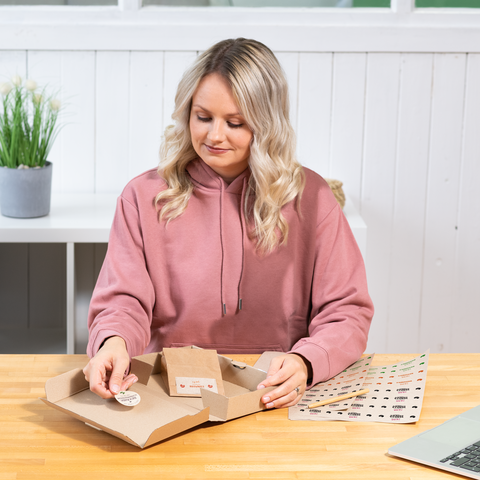 Eine Person mit blonden, lockigen Haaren sitzt an einem Tisch und arbeitet an einer Verpackung, während sie sich Aufkleber anschaut.