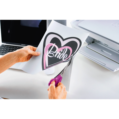 Eine Hand schneidet ein Blatt Papier mit einem Herzmotiv und dem Wort "Bride" aus, während ein Laptop und ein Drucker im Hintergrund sichtbar sind.