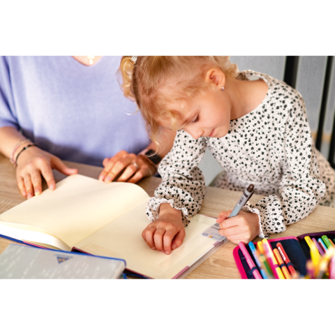 Ein kleines Mädchen schreibt in ein Notizbuch, während eine erwachsene Person daneben sitzt und zuschaut. Bunte Stifte sind auf dem Tisch verteilt.