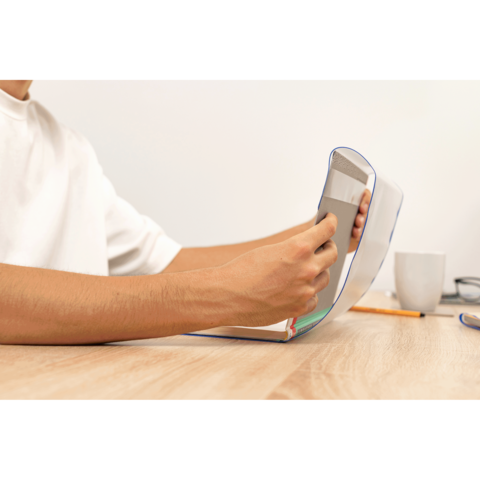 Eine Hand hält ein Smartphone, während die andere eine aufgeschlagene Notizbuchseite berührt. Ein Kaffeebecher und Schreibutensilien sind im Hintergrund sichtbar.