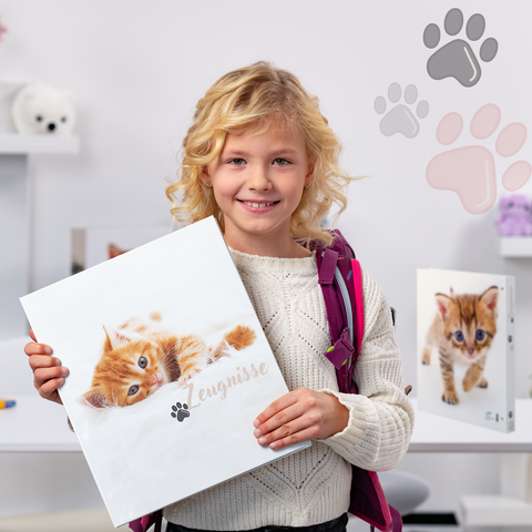 Ein Mädchen mit blonden Locken hält einen Ordner mit einem Katzenbild. Im Hintergrund sind weitere Ordner und Spielzeug zu sehen."
(Note: The text is already in German and does not require translation.)