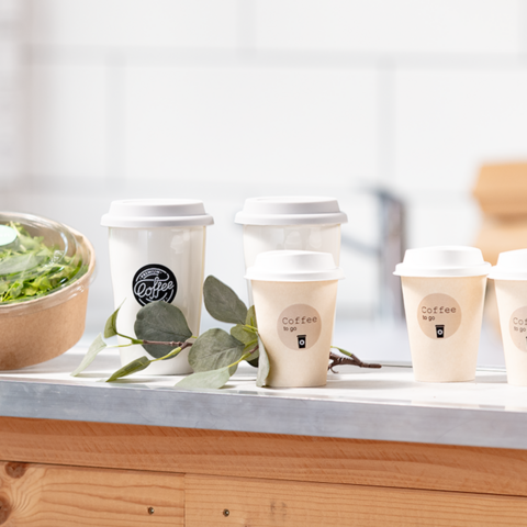 Auf einem Tisch stehen mehrere Kaffeebecher neben einer Schüssel Salat. Die Becher sind mit To-Go-Aufschriften versehen.
