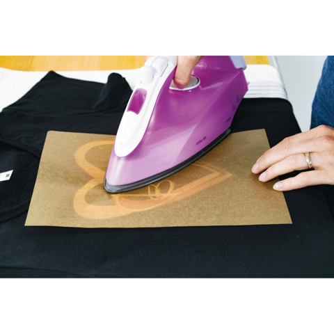 Eine Person bügelt mit einem lila Bügeleisen ein Herzmotiv auf ein schwarzes T-Shirt, das auf einer Tischoberfläche liegt.