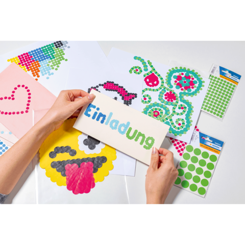 Hände halten eine Einladungskarte mit dem Wort "Einladung", umgeben von bunten Bastelmaterialien und Aufklebern auf einem Tisch.
