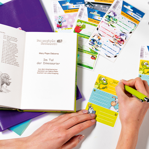 Hände halten eine gelbe Notizkarte mit einem Cartoon-Motiv. Auf einem Tisch liegen ein Buch, Schreibwaren und bunte Aufkleber.