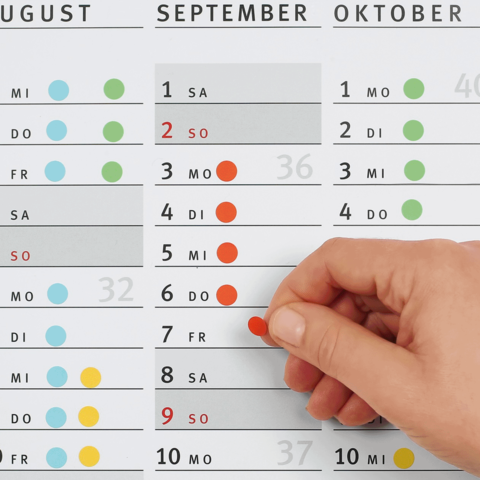 Ein Hand zeigt auf einen Kalender, der die Monate August, September und Oktober mit markierten Daten und Wochentagen anzeigt.