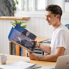 Ein junger Mann sitzt an einem Tisch und betrachtet ein aufgeschlagenes Buch mit Landschaftsbildern. Ein Kaffeebecher steht neben ihm.