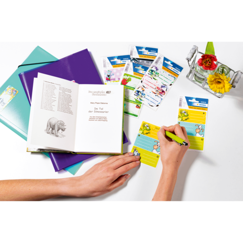 Eine Hand schreibt in ein Notizbuch, während ein offenes Buch mit einem Elefantenbild und mehrere Karten im Hintergrund liegen.