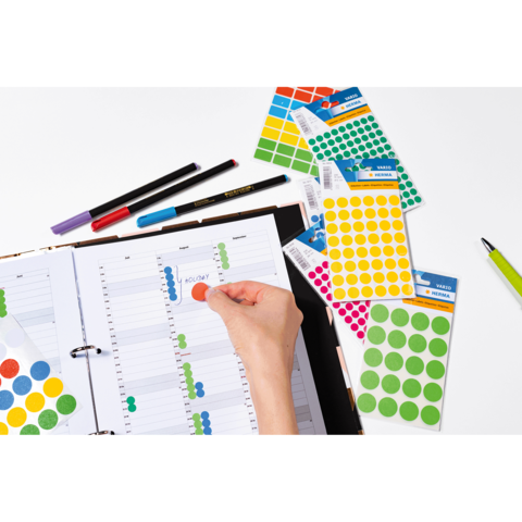 Ein offenes Notizbuch mit einem Kalender, auf dem bunte Aufkleber platziert werden. Daneben liegen Marker und weitere Aufkleberblätter.
