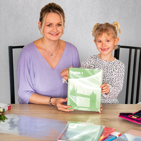 Eine Frau und ein Mädchen sitzen an einem Tisch, beide lächeln und halten ein grünes Heft. Auf dem Tisch liegen Schreibmaterialien und Blumen.