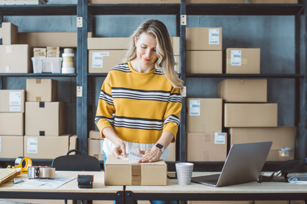 Eine Frau in einem gestreiften Pullover steht an einem Tisch, umgeben von Kartons und einem Laptop in einem modernen Büro." 

(Note: The text is already in German and does not require translation. It is clear and fluent as it is.)
