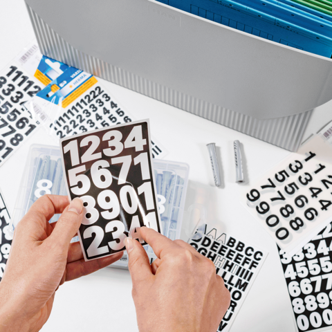 Hände halten eine transparente Box mit Zahlen und Buchstabenaufklebern, umgeben von weiteren Aufklebern und einem Ordner im Hintergrund.