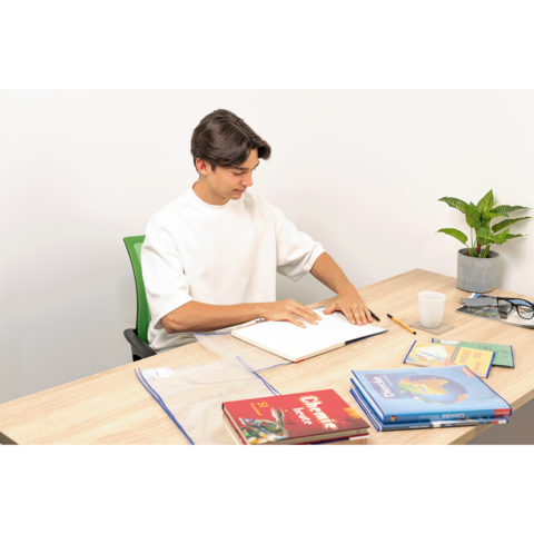 Ein junger Mann sitzt an einem Tisch, blättert in einem Notizbuch und hat mehrere Bücher sowie eine Pflanze und eine Tasse neben sich.