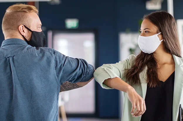 Zwei Personen in einem modernen Büro, die sich mit dem Ellenbogen begrüßen, während sie beide Gesichtsmasken tragen.