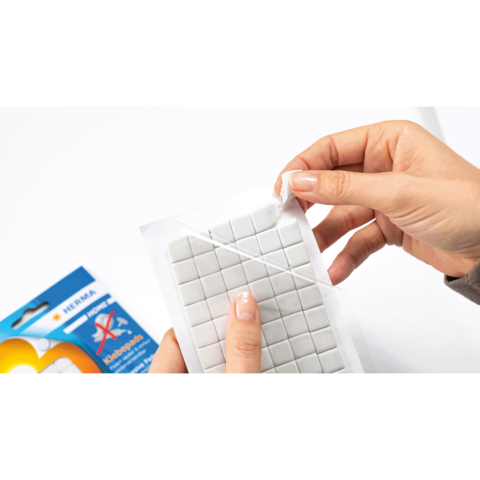 Eine Hand zieht eine transparente Folie von einem Paket mit kleinen, quadratischen, weißen Klebepunkten ab. Im Hintergrund ist eine Verpackung sichtbar.