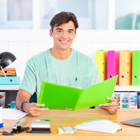 Ein junger Mann sitzt an einem Schreibtisch und hält eine grüne Mappe. Auf dem Tisch sind Schreibwaren und bunte Ordner sichtbar.