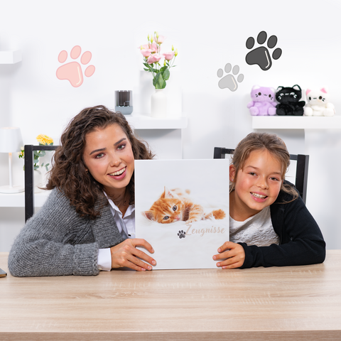 Eine Frau und ein Mädchen sitzen an einem Tisch und halten ein Buch mit einem Kätzchen-Cover. Im Hintergrund sind dekorative Elemente zu sehen.