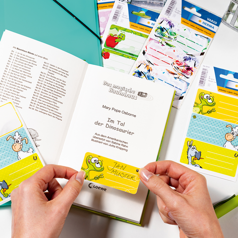 Hände halten eine bunte Notizkarte mit einem Cartoon-Motiv, umgeben von Schulmaterialien, einem Buch und einer Vase mit Blumen.
