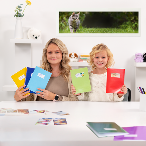 Eine Frau und ein Mädchen halten bunte Ordner in der Hand, während sie an einem Tisch mit Fotos sitzen. Im Hintergrund ist eine Katze zu sehen.