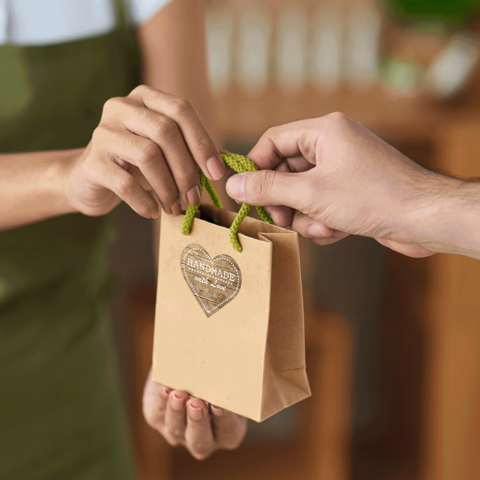 Eine Person übergibt eine kleine, braune Papiertüte mit einem Herzanhänger an eine andere Person. Die Tüte ist als handgemacht gekennzeichnet.