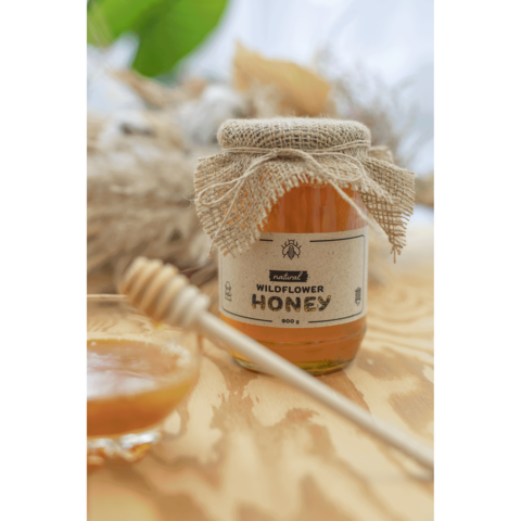 Glas Honig mit Jute-Deckel und Etikett "Wildflower Honey", daneben ein Honiglöffel und eine kleine Schale mit Honig.