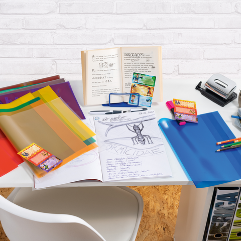 Bunte Kunststoffmappen, ein Stapel Notizen und ein Hefter liegen auf einem Tisch. Im Hintergrund steht ein offenes Buch mit handschriftlichen Notizen.