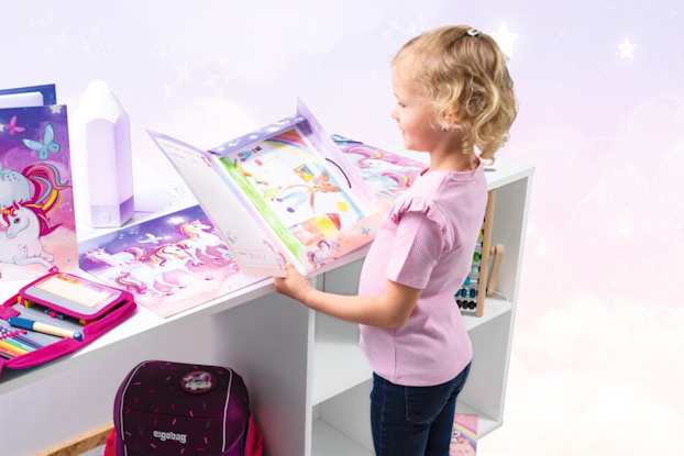 Ein Mädchen steht an einem Tisch und schaut sich ein buntes Buch mit Einhornmotiven an. Neben ihr liegen weitere Bücher und ein Rucksack.