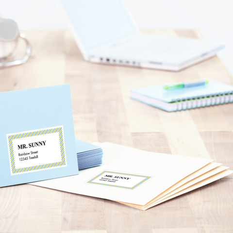 Blaue und weiße Briefumschläge mit Adressetiketten liegen auf einem Tisch, neben einem Laptop und einem Notizbuch." 

(Note: The text is already in German and does not require translation. It is clear and fluent as is.)