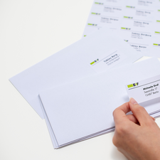 Ein junger Mann sitzt an einem Tisch, lächelt und hält ein Blatt mit Aufklebern in der Hand, während er vor einem Laptop und einer Verpackung steht." 

(Note: The text is already in German and does not require translation. It is clear and fluent as it is.)