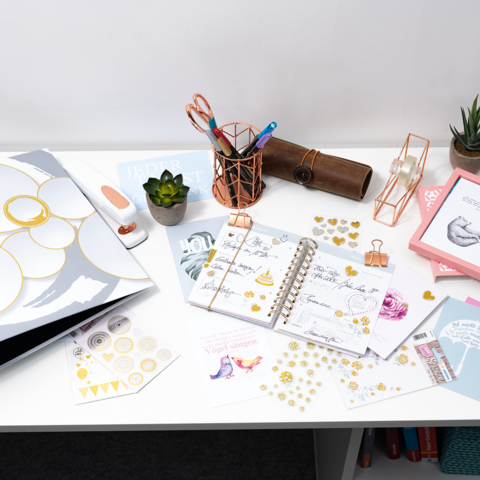 Schreibtisch mit Planer, Schreibutensilien, einer Pflanze und verschiedenen Dekorationselementen. Farbiges Papier und Aufkleber sind verteilt.
