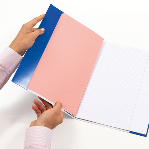 Eine Person hält ein geöffnetes Notizbuch mit einem blauen und rosa Einband, das leere, linierte Seiten zeigt.