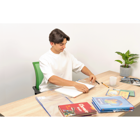 Ein junger Mann sitzt an einem Tisch, blättert in einem Notizbuch und hat Bücher sowie Schreibmaterialien vor sich. Eine Pflanze ist im Hintergrund.