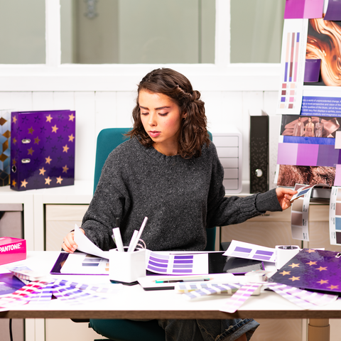 Eine Person sitzt an einem Tisch, umgeben von Farbmustern und Designmaterialien, während sie an einem kreativen Projekt arbeitet." 

(Note: The text is already in German and does not require translation. It is clear and fluent as it is.)