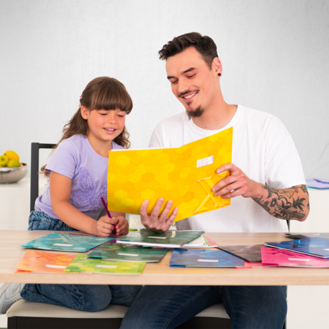 Ein Mann und ein Mädchen sitzen an einem Tisch, während sie an einem Projekt arbeiten. Der Mann hält eine gelbe Mappe, das Mädchen zeichnet.