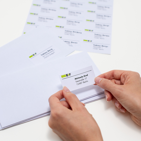 Ein junger Mann sitzt an einem Tisch, lächelt und hält ein Blatt mit Aufklebern in der Hand. Vor ihm steht ein Karton und ein Laptop."
(Note: The text is already in German and does not require translation. It is clear and fluent as it is.)