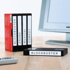 Eine Sammlung von VHS-Kassetten mit den Beschriftungen "BLOCKBUSTER", "DRAMA", "MUSICAL" und "COMEDY" neben einer Fernbedienung und einem Computer.