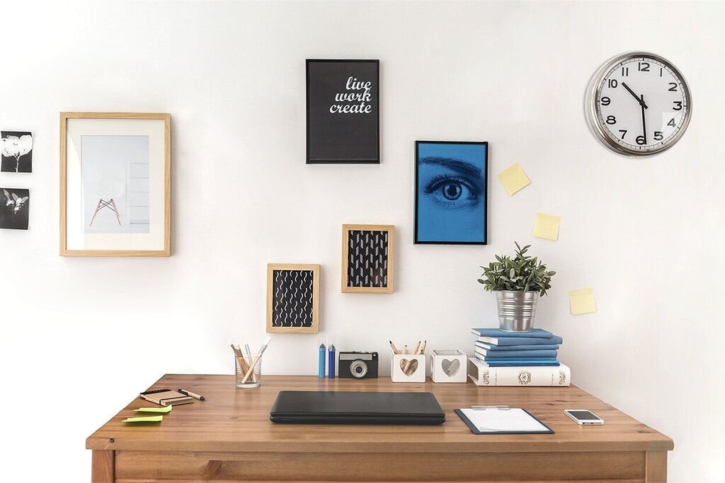 Ein Schreibtisch mit einem Laptop, dekorativen Bildern an der Wand, einer Pflanze und einer Stehlampe. Eine Wanduhr zeigt die Uhrzeit an.