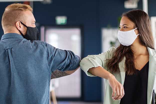 Zwei Personen in einem modernen Büro, die sich mit dem Ellenbogen begrüßen, während sie beide Gesichtsmasken tragen.