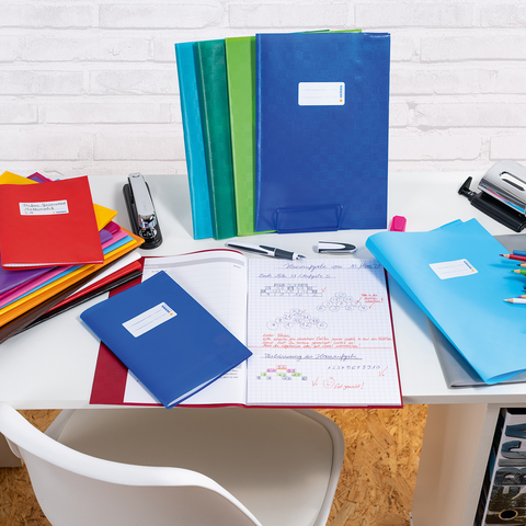 Bunte Ordner und Hefte auf einem Schreibtisch, daneben ein Stift und ein Hefter. Ein offenes Heft mit handschriftlichen Notizen liegt bereit.