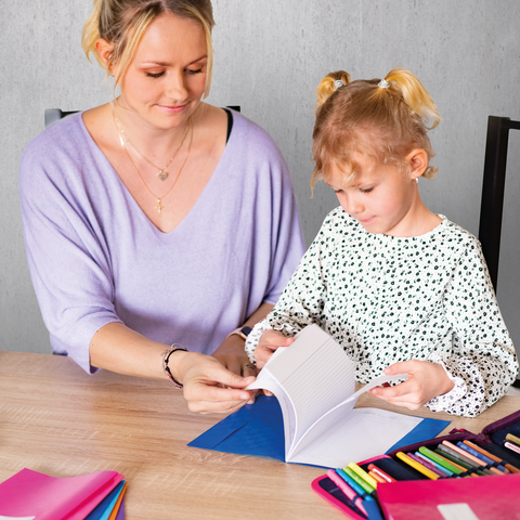 Eine Frau und ein kleines Mädchen sitzen an einem Tisch, während sie ein Buch durchblättern. Bunte Stifte und Notizblätter liegen auf dem Tisch.