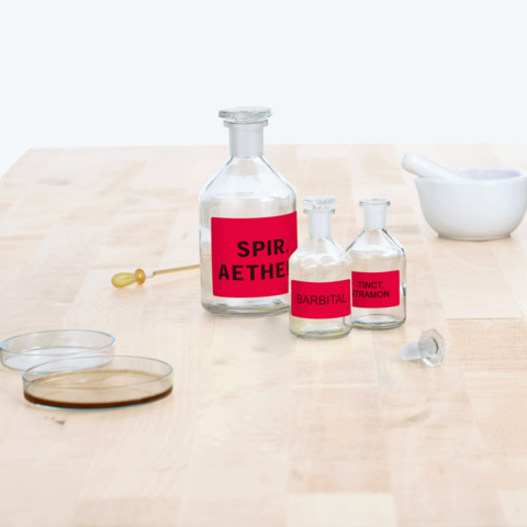 Flaschen mit roten Etiketten, beschriftet mit "Spir. Aether.", "Barbital" und "Tinct. Tramon", sowie ein Mörser und einige Glasplatten auf einem Tisch.