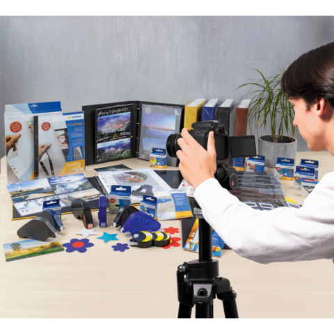 Eine Person fotografiert eine Vielzahl von Büro- und Bastelmaterialien, darunter Ordner, Kleber und Bastelwerkzeuge, auf einem Tisch.