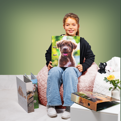 Ein Mädchen sitzt auf einem Pouf und hält einen Ordner mit einem Bild von einem Hund. Neben ihr liegen weitere Ordner und eine kleine Pflanze.